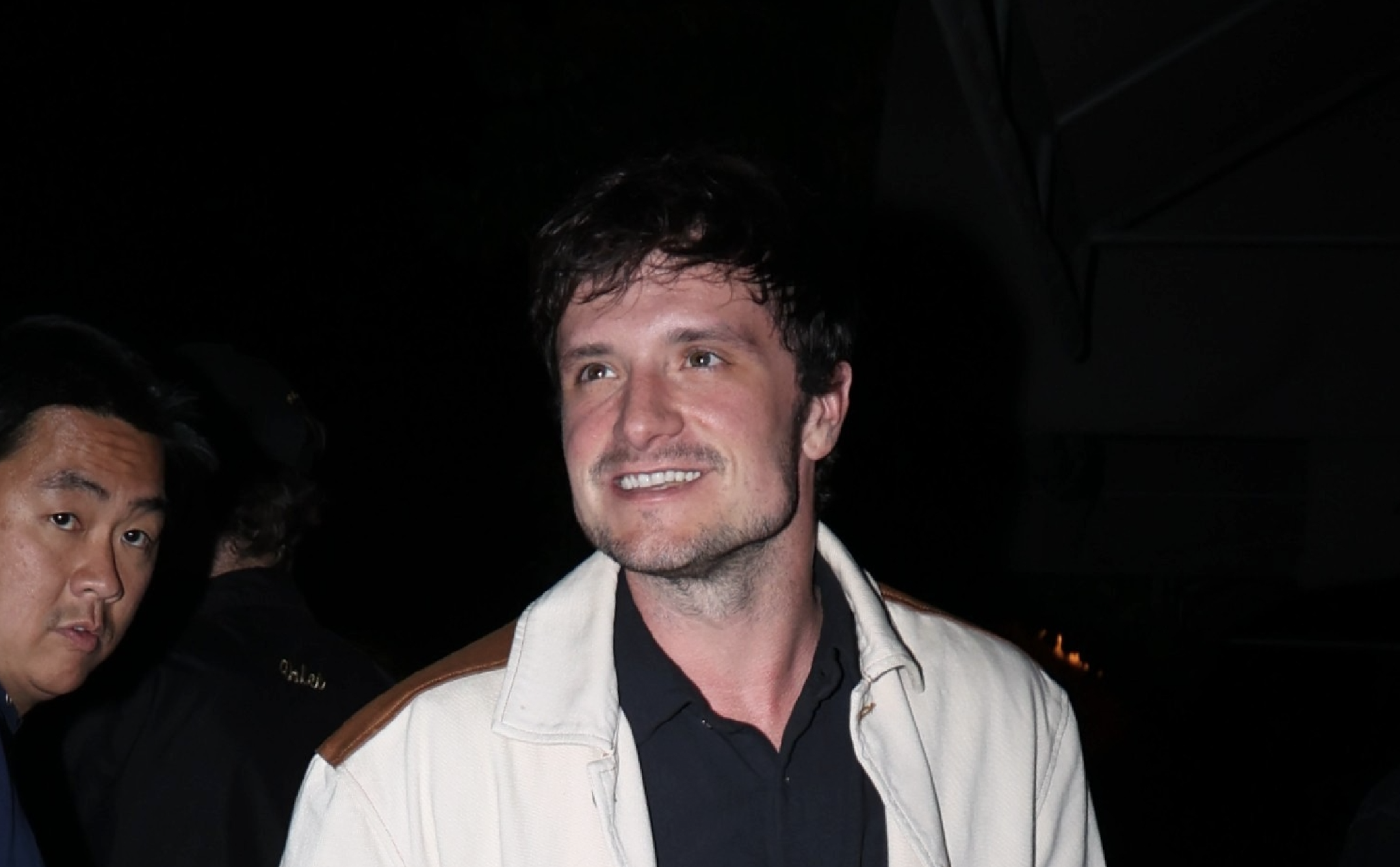 Josh Hutcherson, wearing a navy blue collared shirt under a white leather jacket looks off as he exits a car in a paparazzi photo. He has scruff and messy dark brown hair.